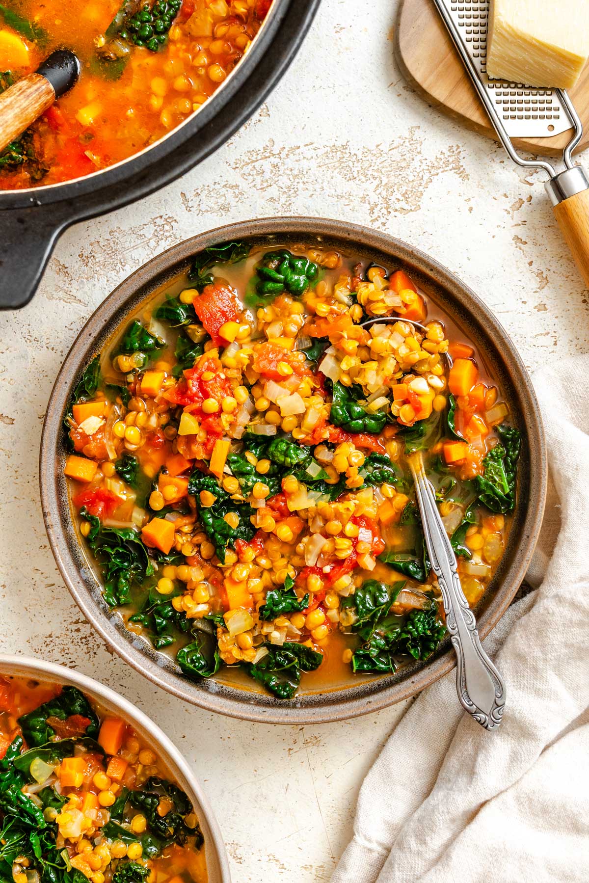 A bowl of lentil soup filled with greens, tomatoes and vegetables with a spoon resting inside.