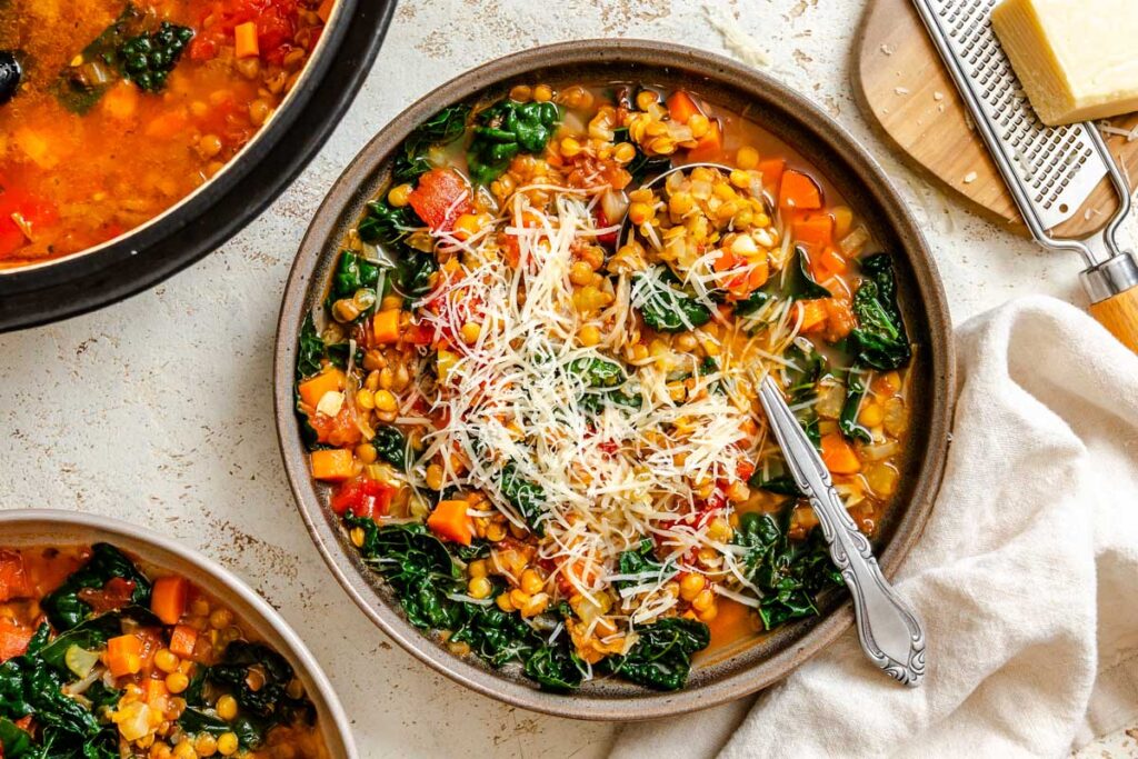 A bowl of lentil soup next to the pot, with tender lentils, kale and tomatoes visible throughout.