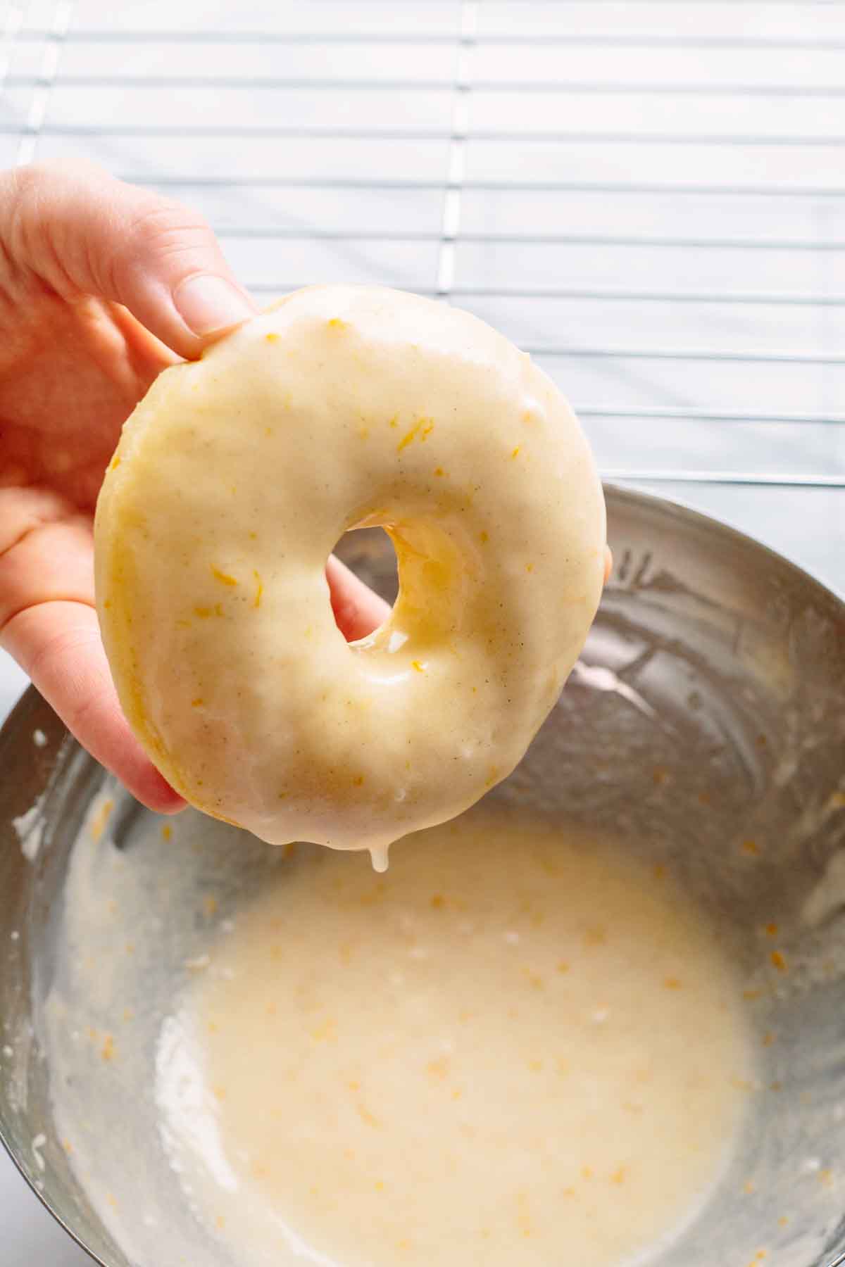 A hand holding a freshly baked donut coated in Meyer lemon glaze over a metal bowl filled with citrus glaze.