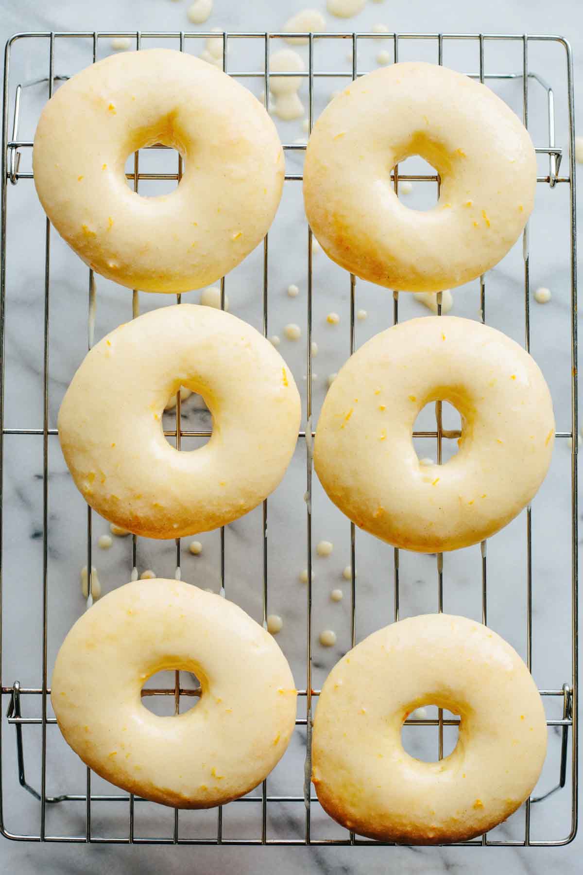 Six pale, freshly glazed Meyer lemon donuts resting on a wire rack with drips of glaze on the surface below.