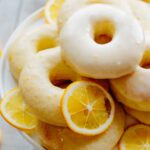 A pile of glazed Meyer lemon donuts on a white plate, tucked in with thin slices of fresh Meyer lemons.