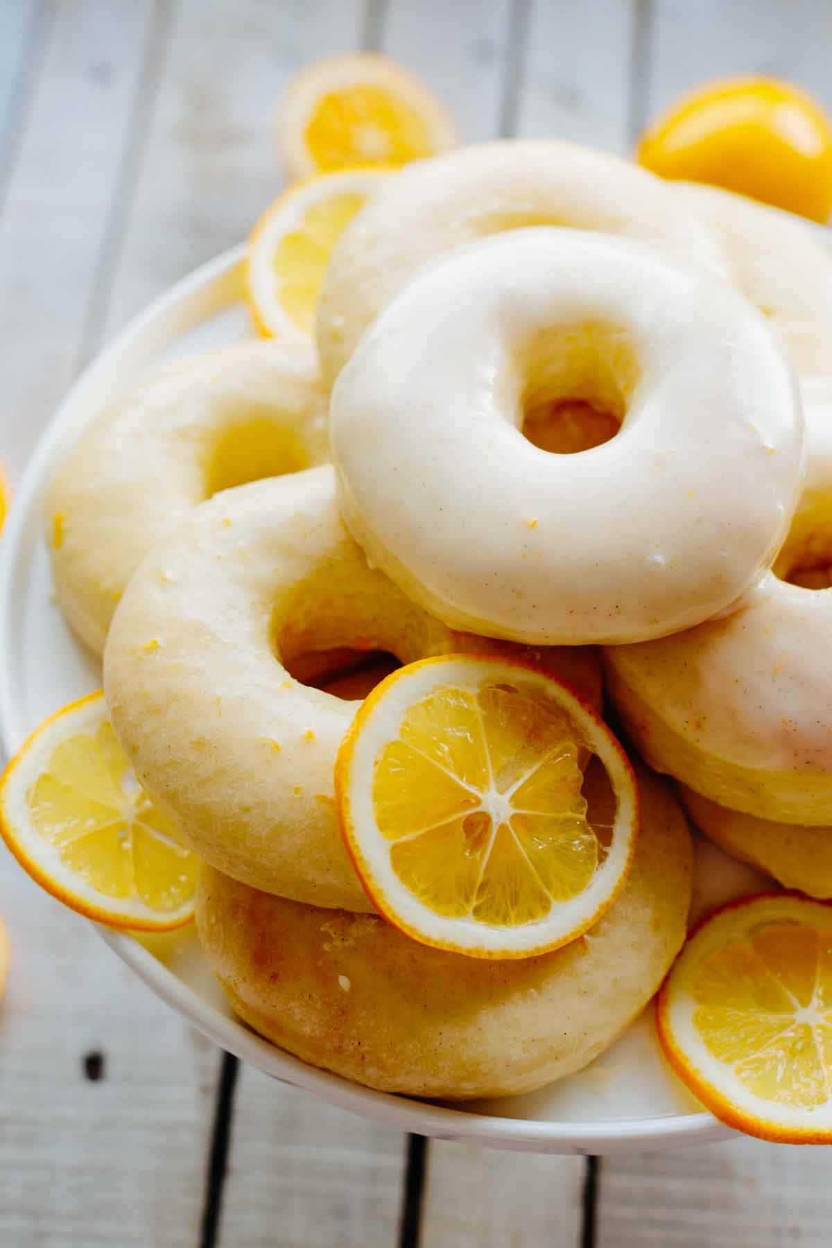 A pile of glazed Meyer lemon donuts on a white plate, tucked in with thin slices of fresh Meyer lemons.