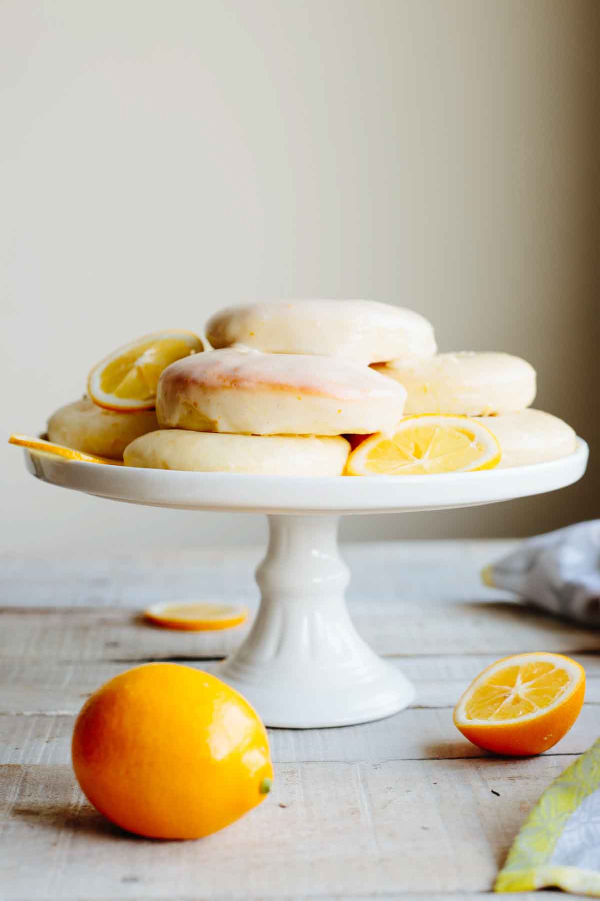 A stack of glazed Meyer lemon donuts arranged on a white pedestal cake stand with whole and sliced Meyer lemons scattered around.