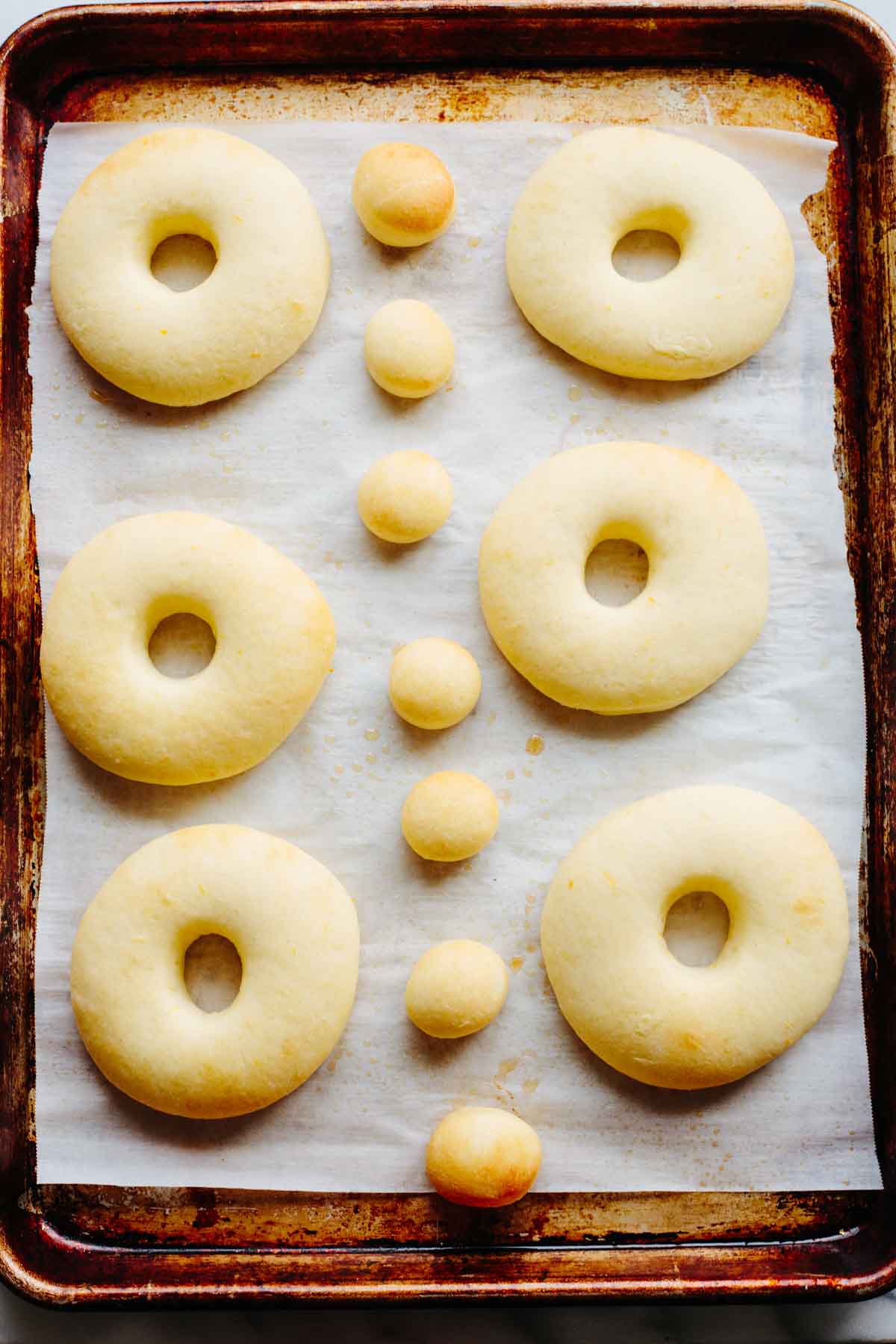 Plain, unglazed donut rings and small donut holes arranged on a parchment lined sheet pan after baking.