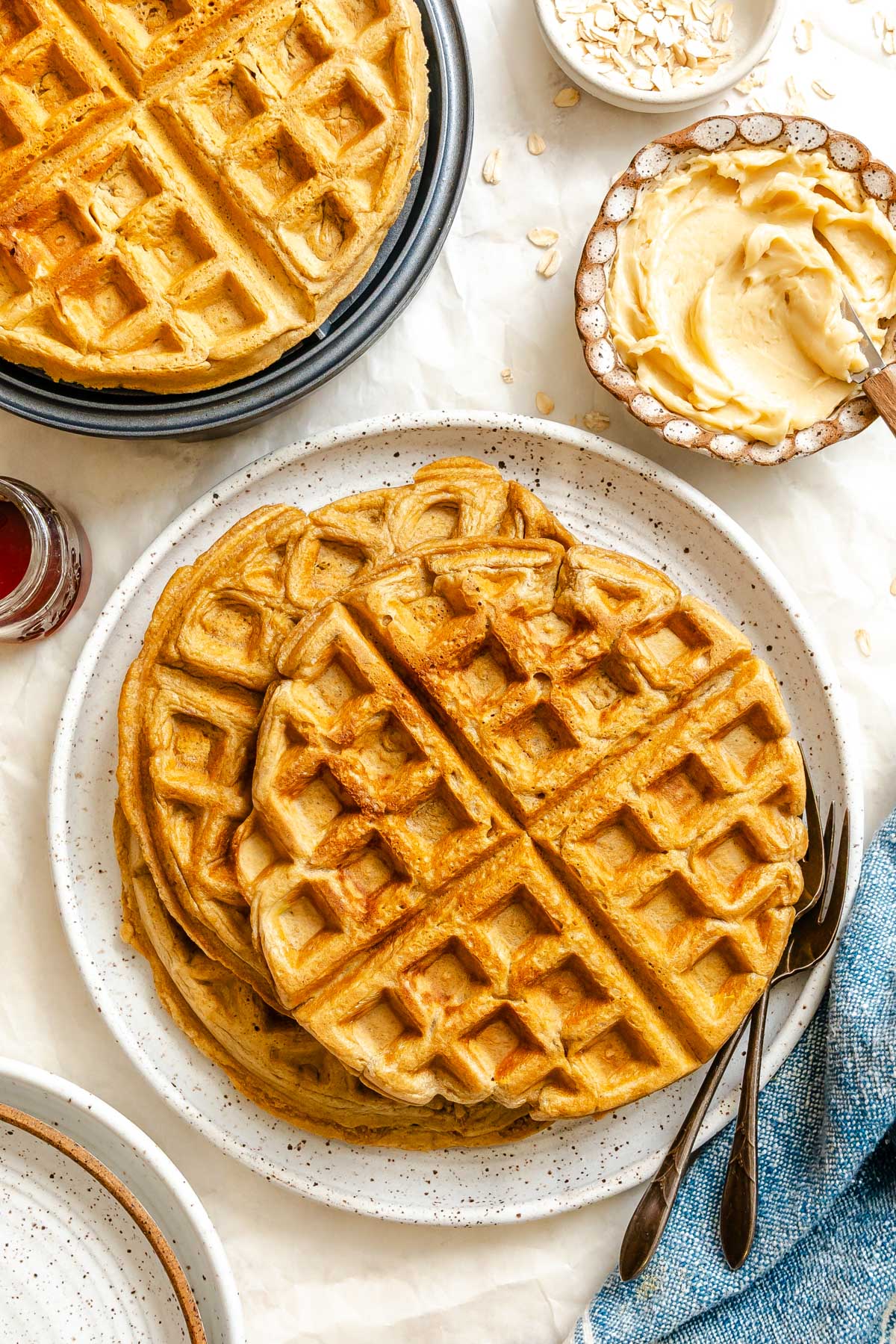 Golden oatmeal waffles stacked on a speckled plate with butter, maple syrup, and oats scattered on a light surface.
