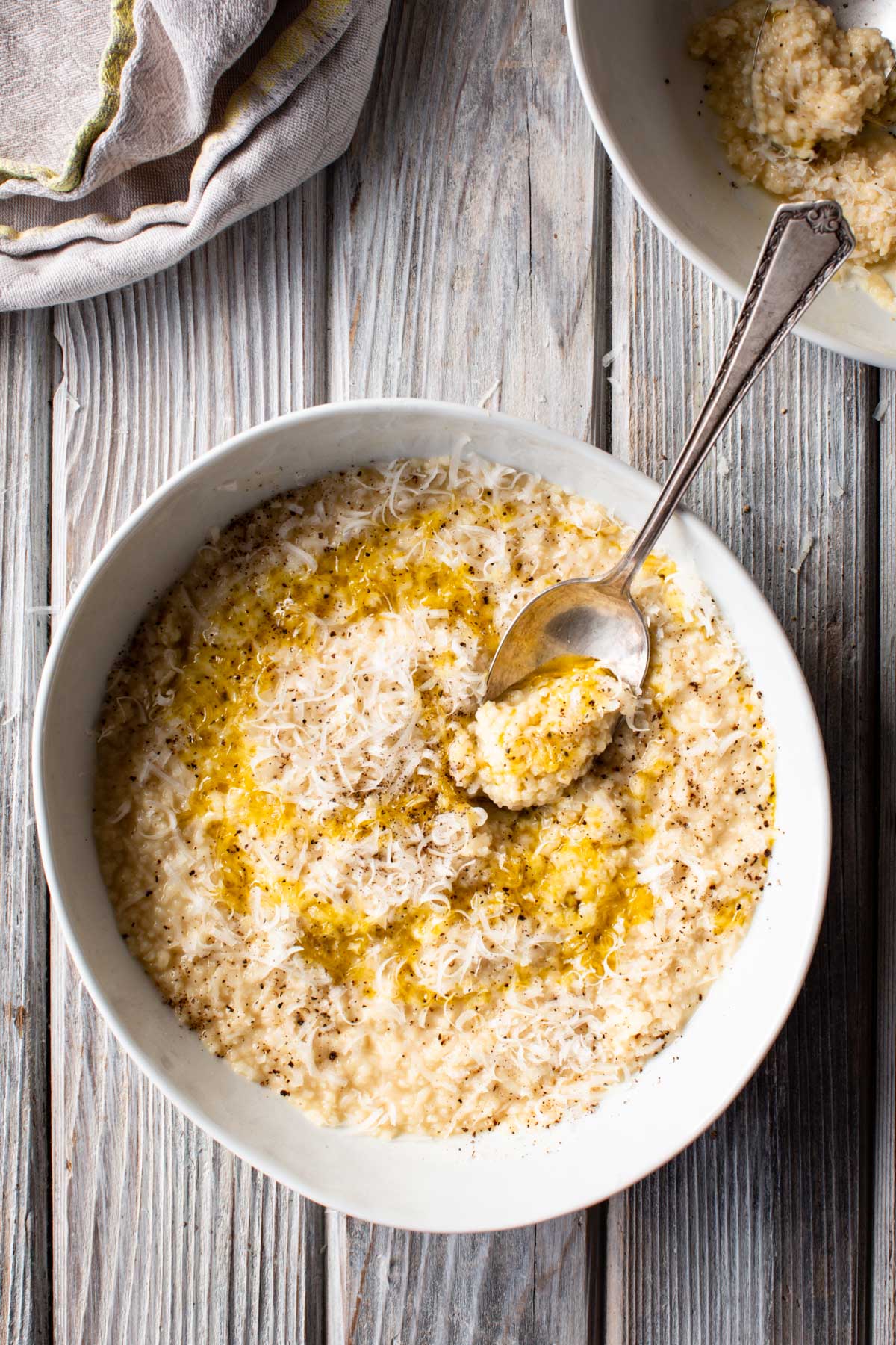 A shallow bowl filled with creamy pastina topped with grated Parmesan, cracked black pepper and a drizzle of olive oil, with a spoon scooping up a bite.