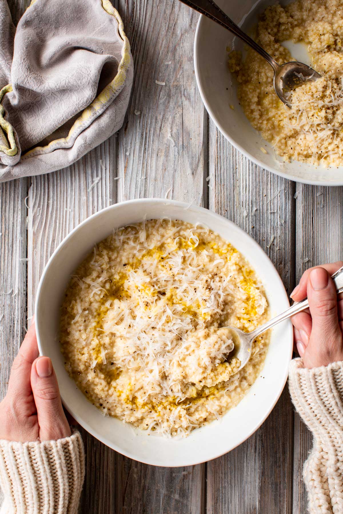 A warm bowl of pastina held between two hands, topped with grated cheese, pepper and olive oil, with another bowl of pastina in the background.