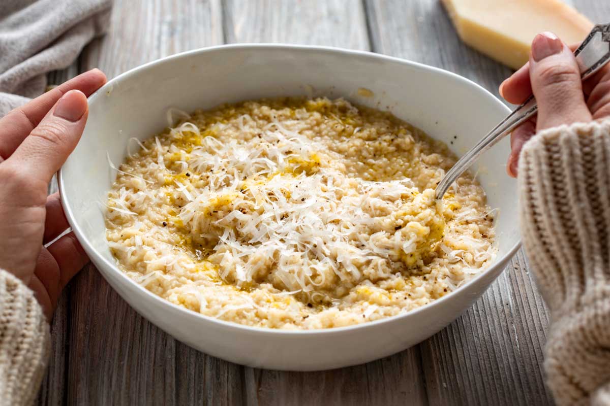 A wide shallow bowl of pastina sits on a rustic wood surface, topped with cheese and olive oil, with hands on both sides and a spoon ready to scoop.