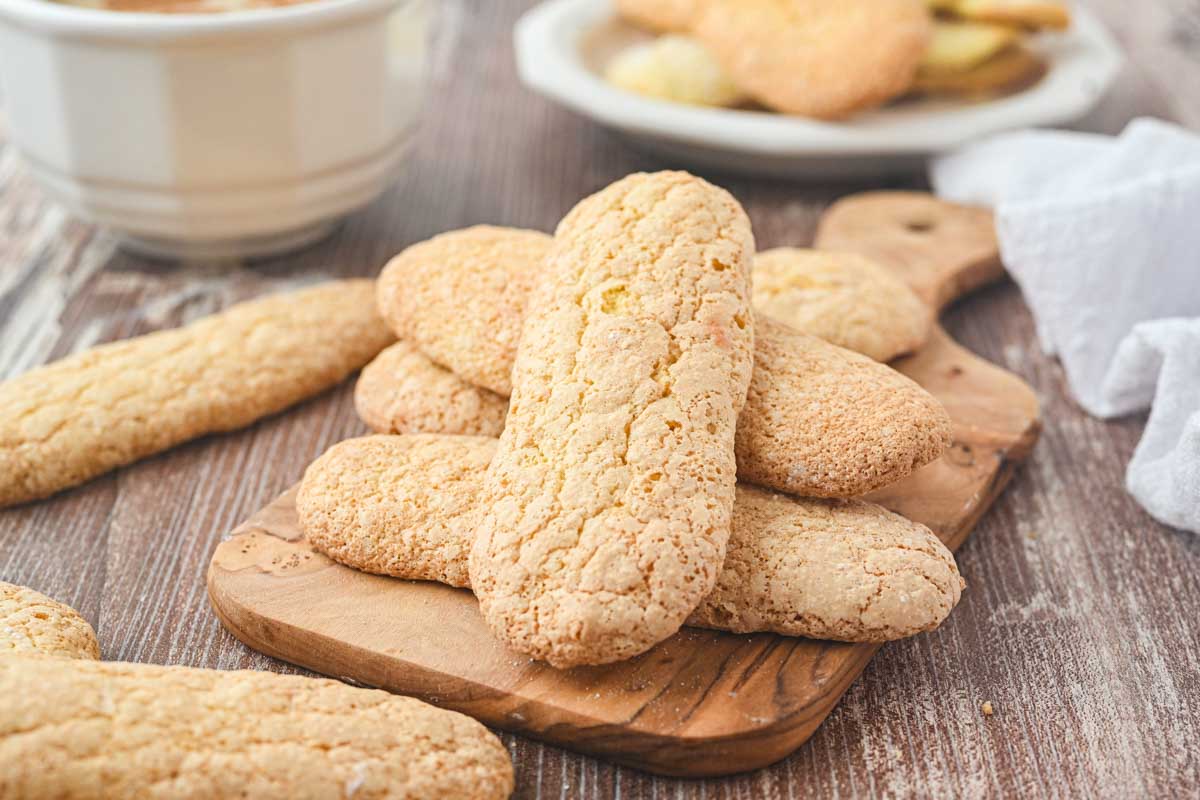 A small stack of crisp, golden ladyfingers displayed on a wooden board with more cookies in the background.