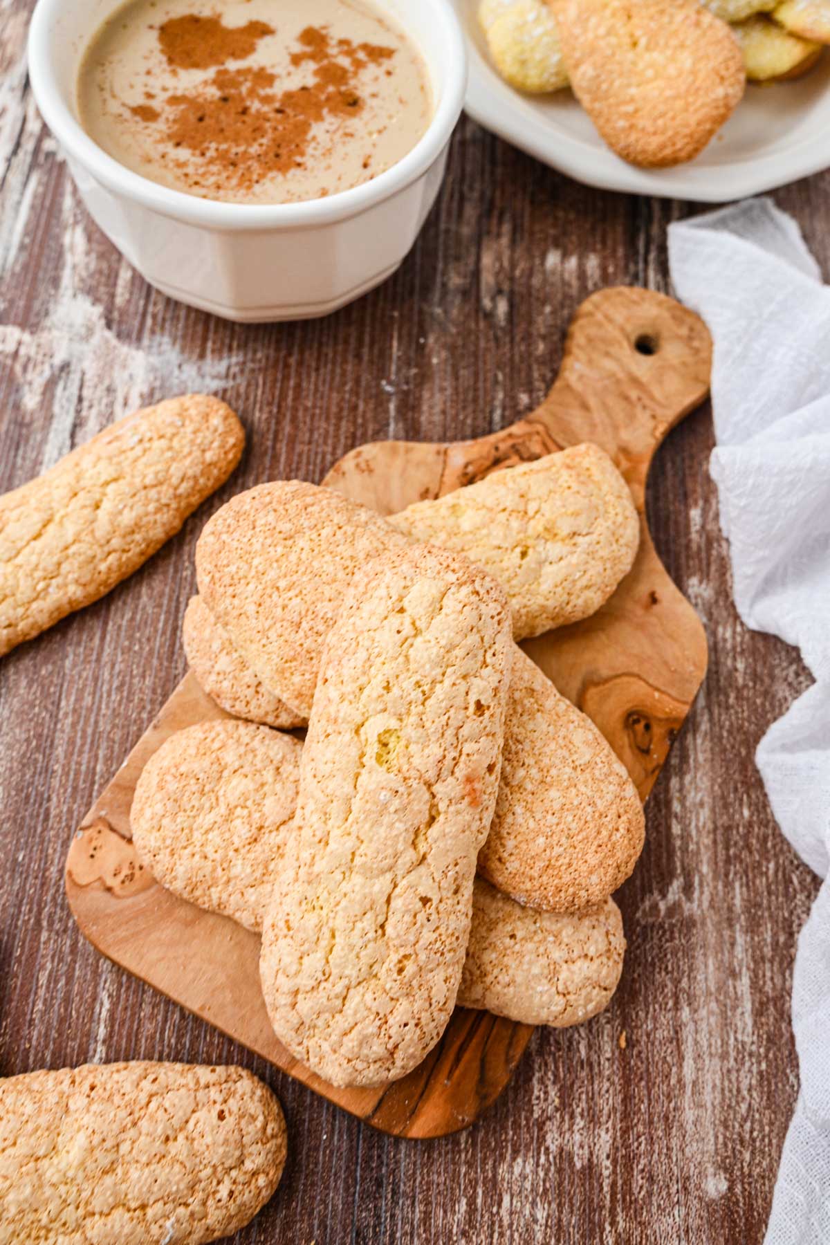 Ladyfingers on a wooden board next to a cup of coffee dusted with cinnamon and a plate of additional cookies.