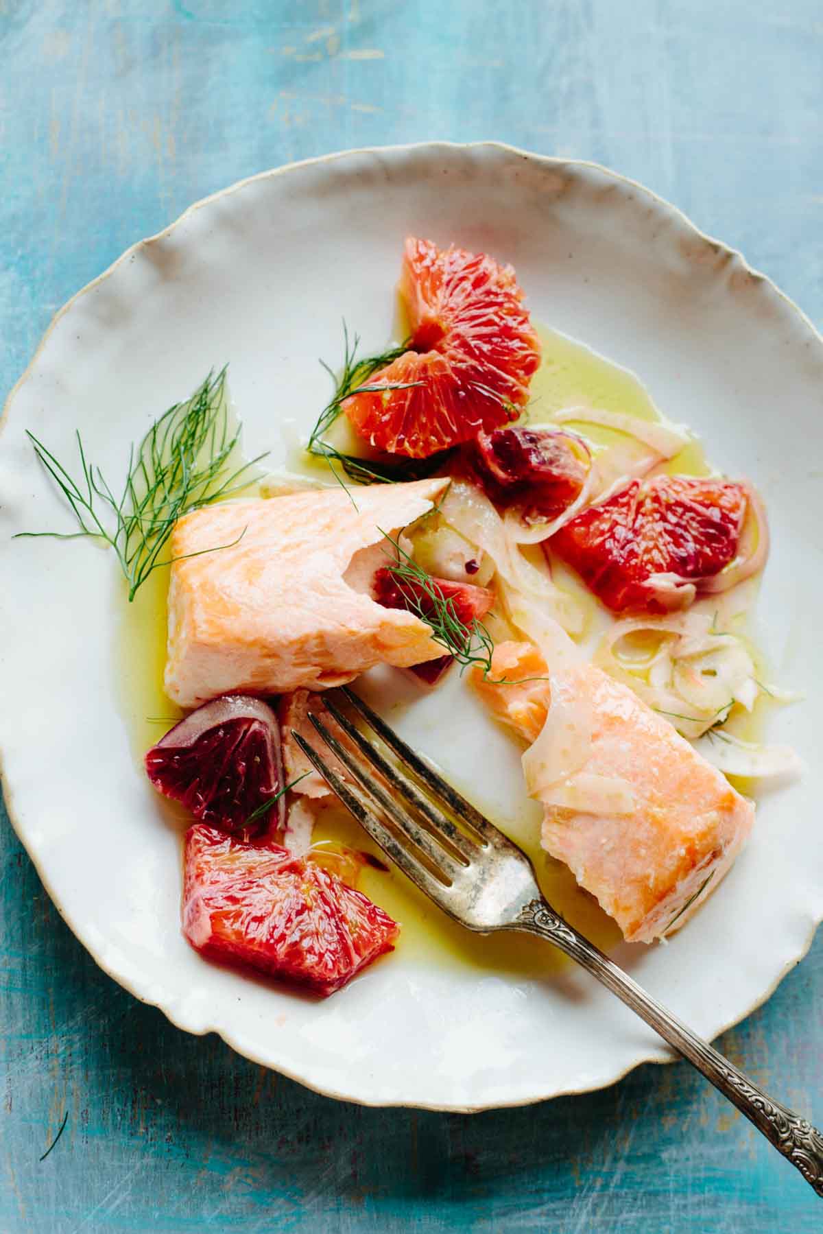 A plated serving of tender slow roasted salmon with shaved fennel, blood orange segments and olive oil, styled with a fork on a white dish.
