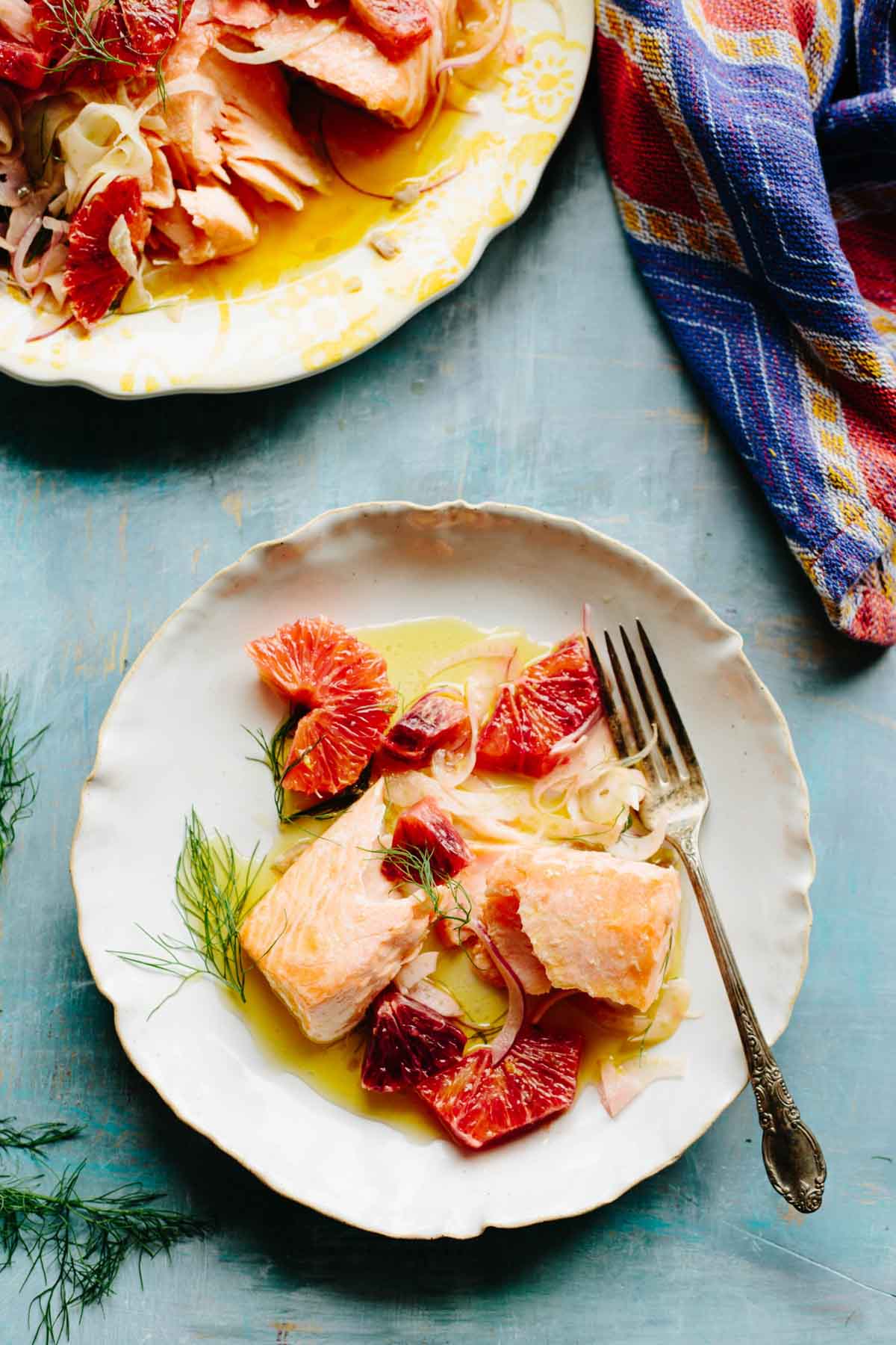 Two plates filled with slow roasted salmon, blood oranges and fennel on a blue surface with a colorful towel alongside.