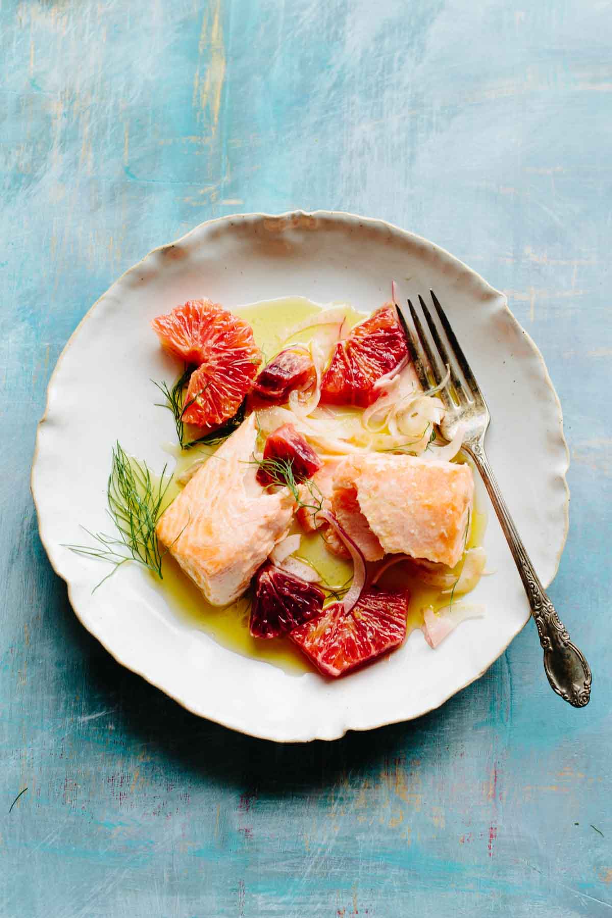 A close up of a plate of roasted salmon with blood orange pieces, thin fennel slices and olive oil on a blue painted background.