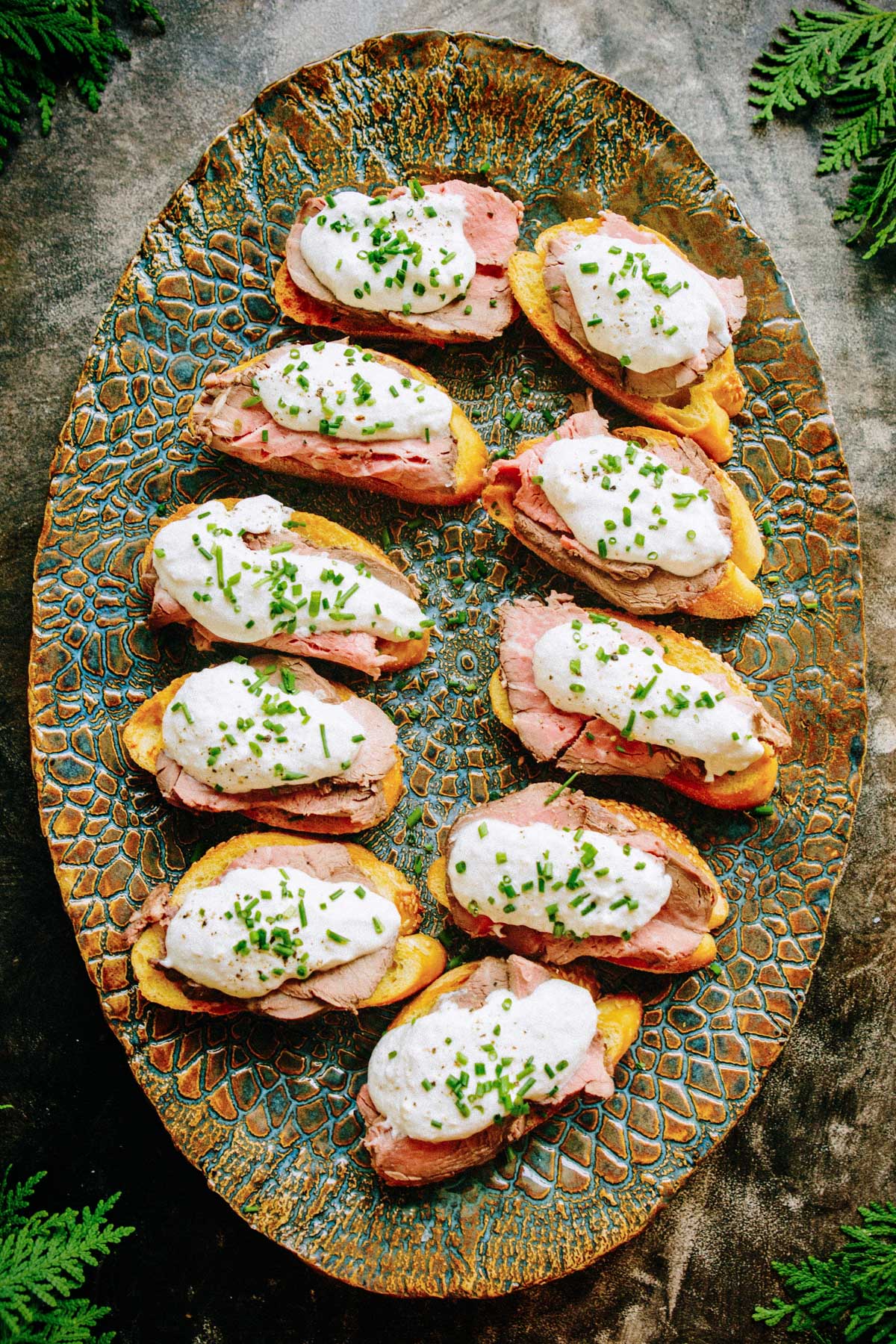 An oval platter of steak crostini topped with horseradish sauce and chives, styled with greenery on a dark background.