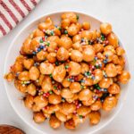 A mound of honey coated struffoli piled high on a white plate and topped with colorful sprinkles.