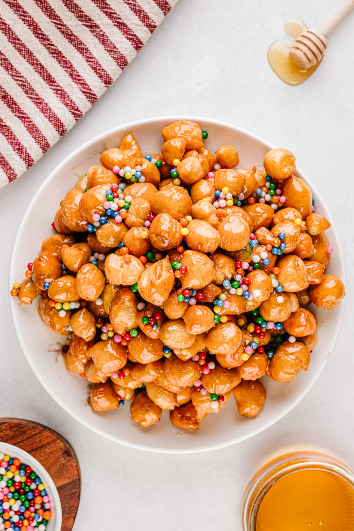 A mound of honey coated struffoli piled high on a white plate and topped with colorful sprinkles.