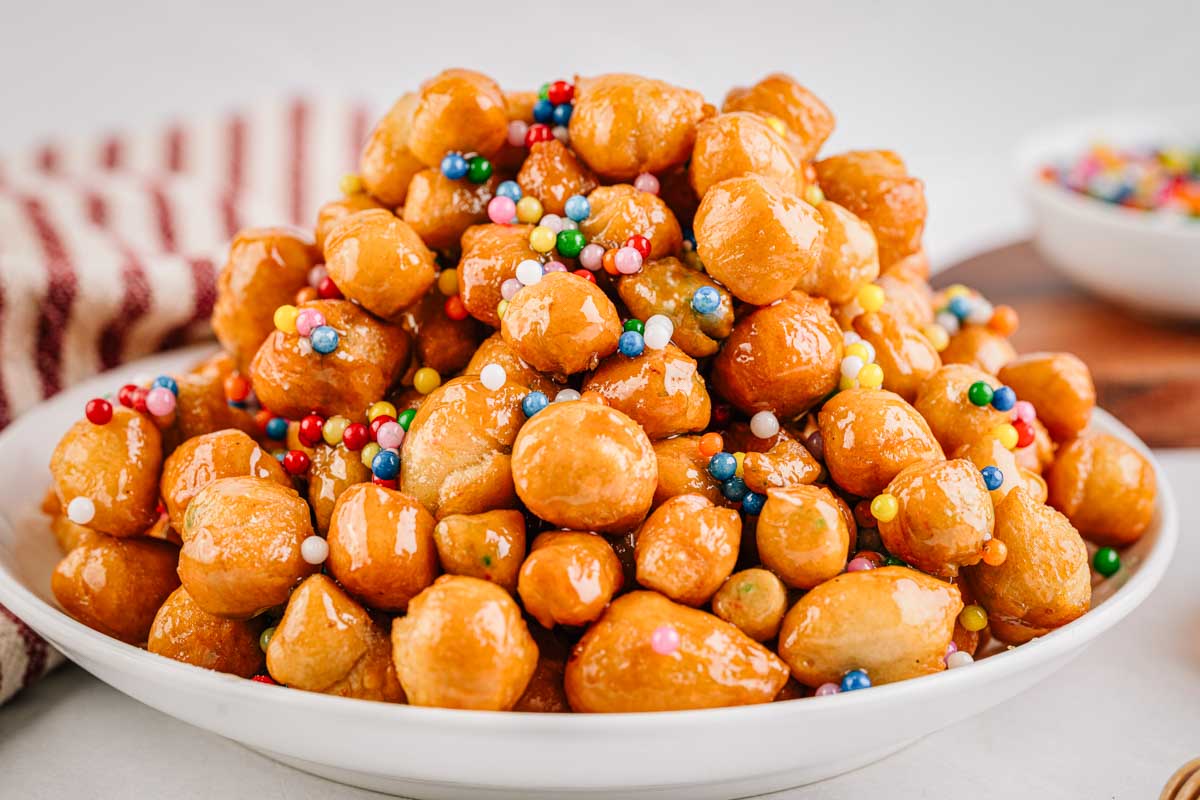 A plate heaped with glossy pignolata, sprinkles scattered throughout, with citrus and a striped towel blurred in the background.