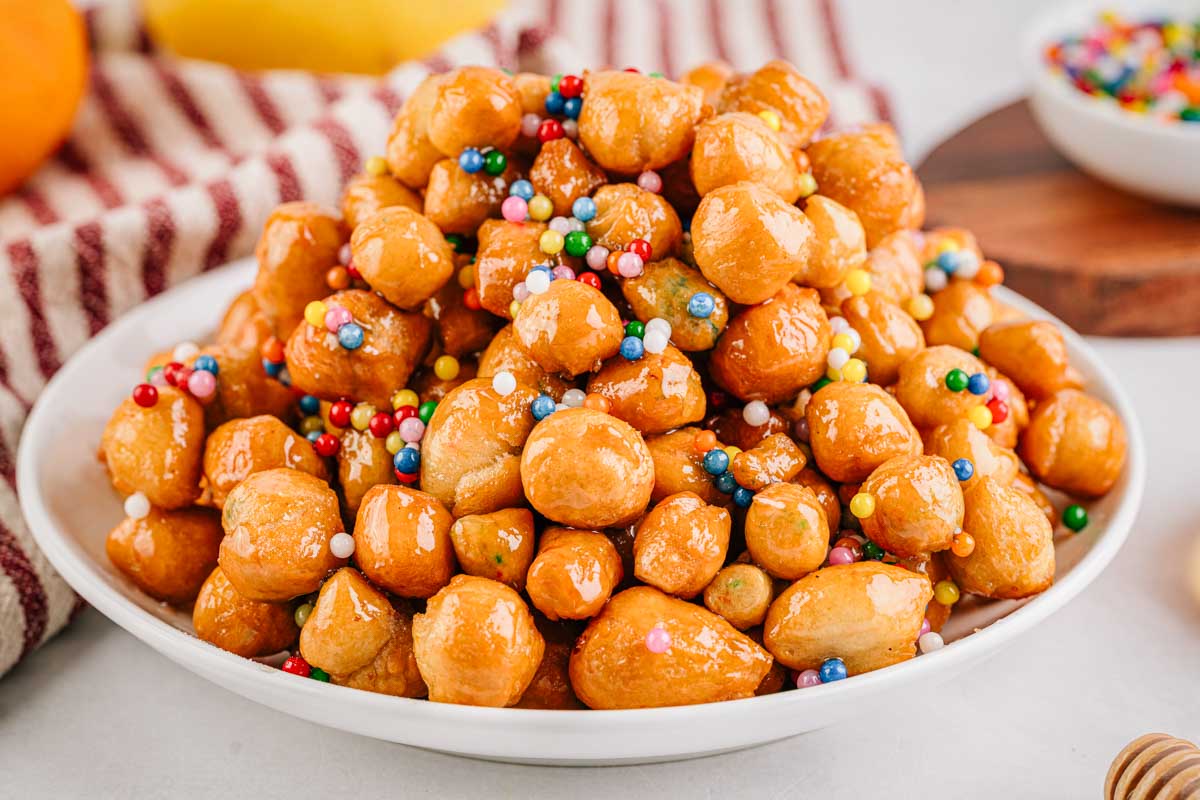 A side angle of the full platter of honey coated struffoli emphasizing their shape, shine and colorful decorations.