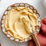 Overhead view of whipped maple butter in a decorative bowl with a knife, maple syrup nearby, and a linen napkin.
