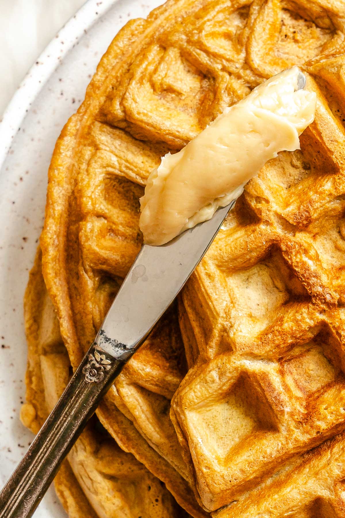 A knife spreading whipped maple butter over a golden waffle, highlighting the soft texture as it melts slightly.