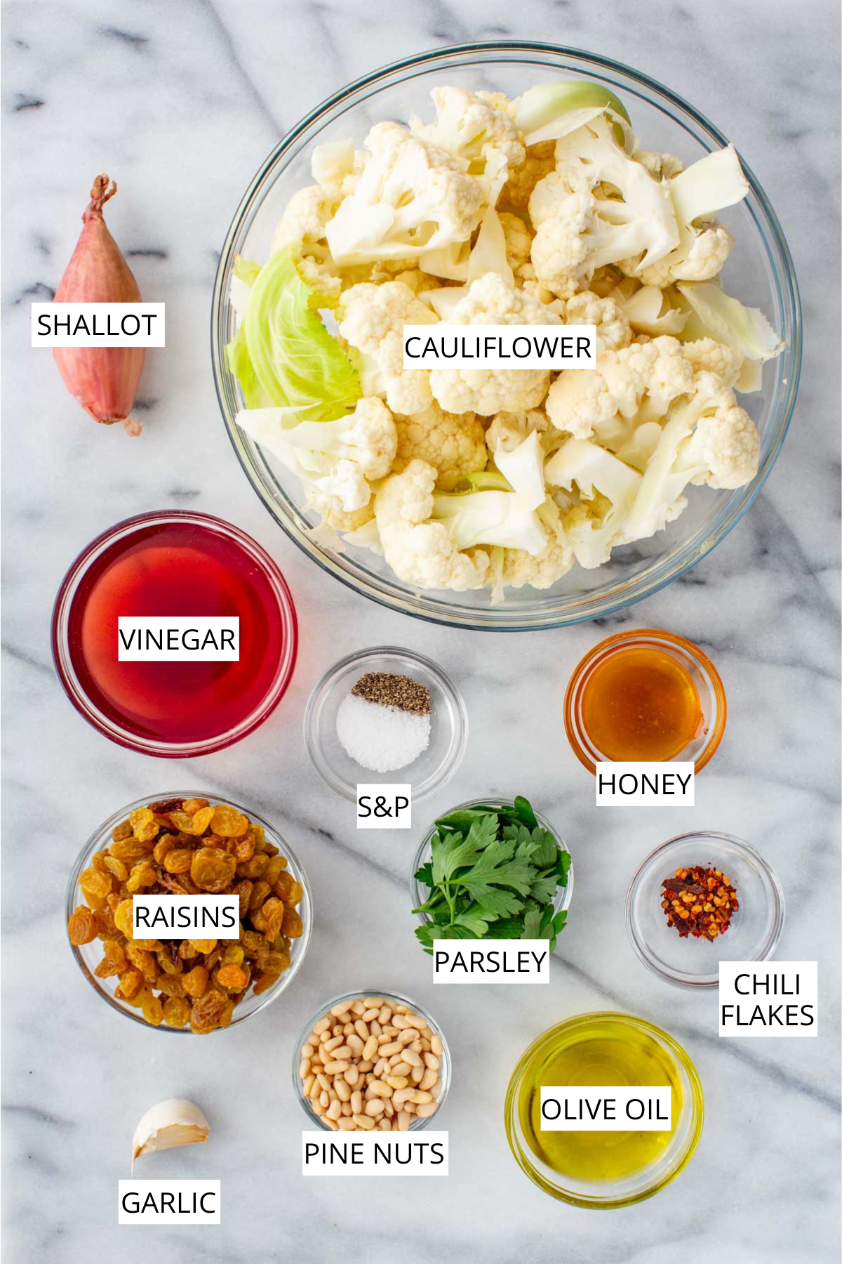 Overhead view of labeled ingredients for cauliflower agrodolce including cauliflower florets, shallot, vinegar, honey, olive oil, golden raisins, pine nuts, parsley, garlic, salt and pepper, and red chili flakes on a marble surface.