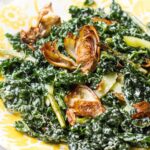 A plated kale salad with golden fried artichoke hearts, roasted asparagus pieces, and pickled celery, generously coated in a creamy parmesan dressing on a white and yellow dish.