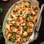Overhead view of cauliflower agrodolce on a platter set on a rustic wooden table with pine nuts and chili flakes nearby.