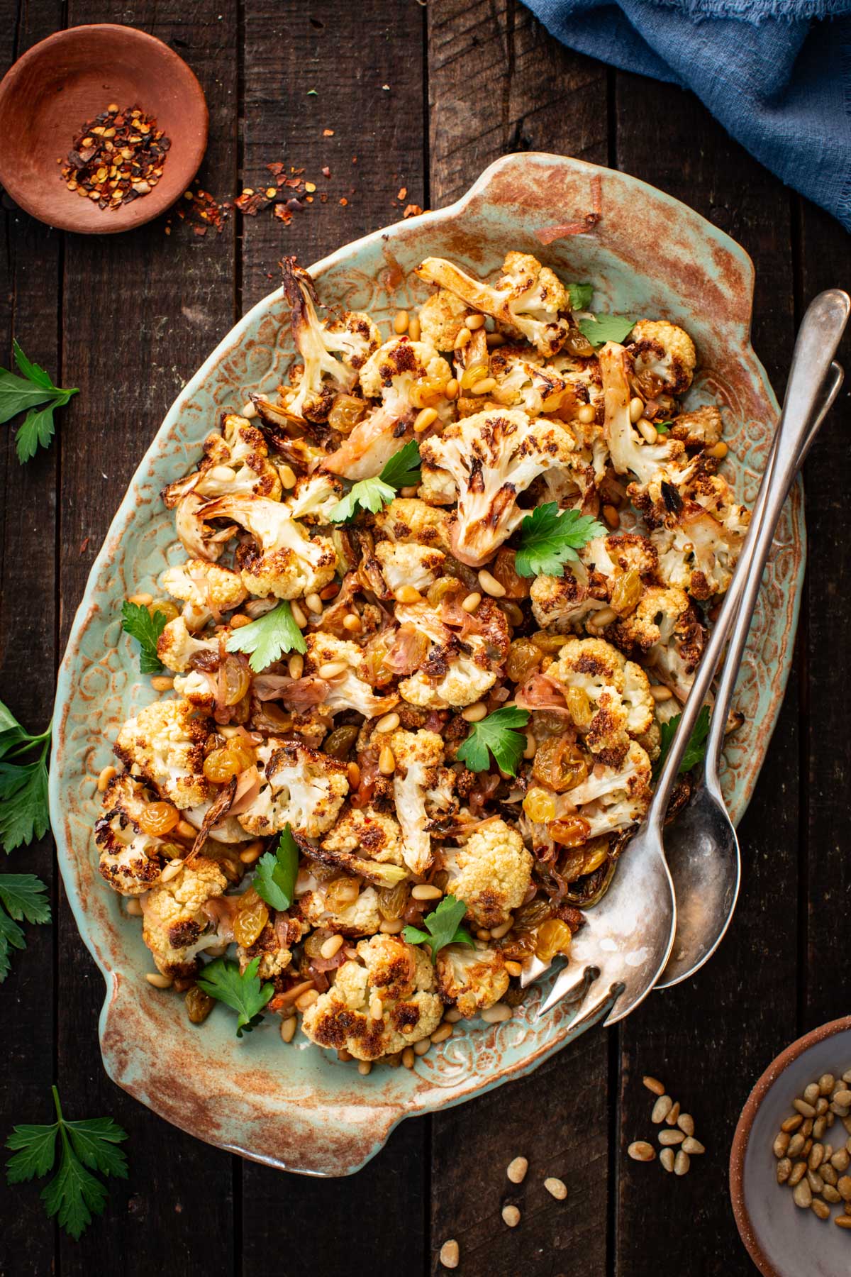 Overhead view of cauliflower agrodolce on a platter set on a rustic wooden table with pine nuts and chili flakes nearby.