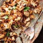 Roasted cauliflower agrodolce tossed with golden raisins, toasted pine nuts, and parsley on a rustic ceramic platter with a serving spoon.