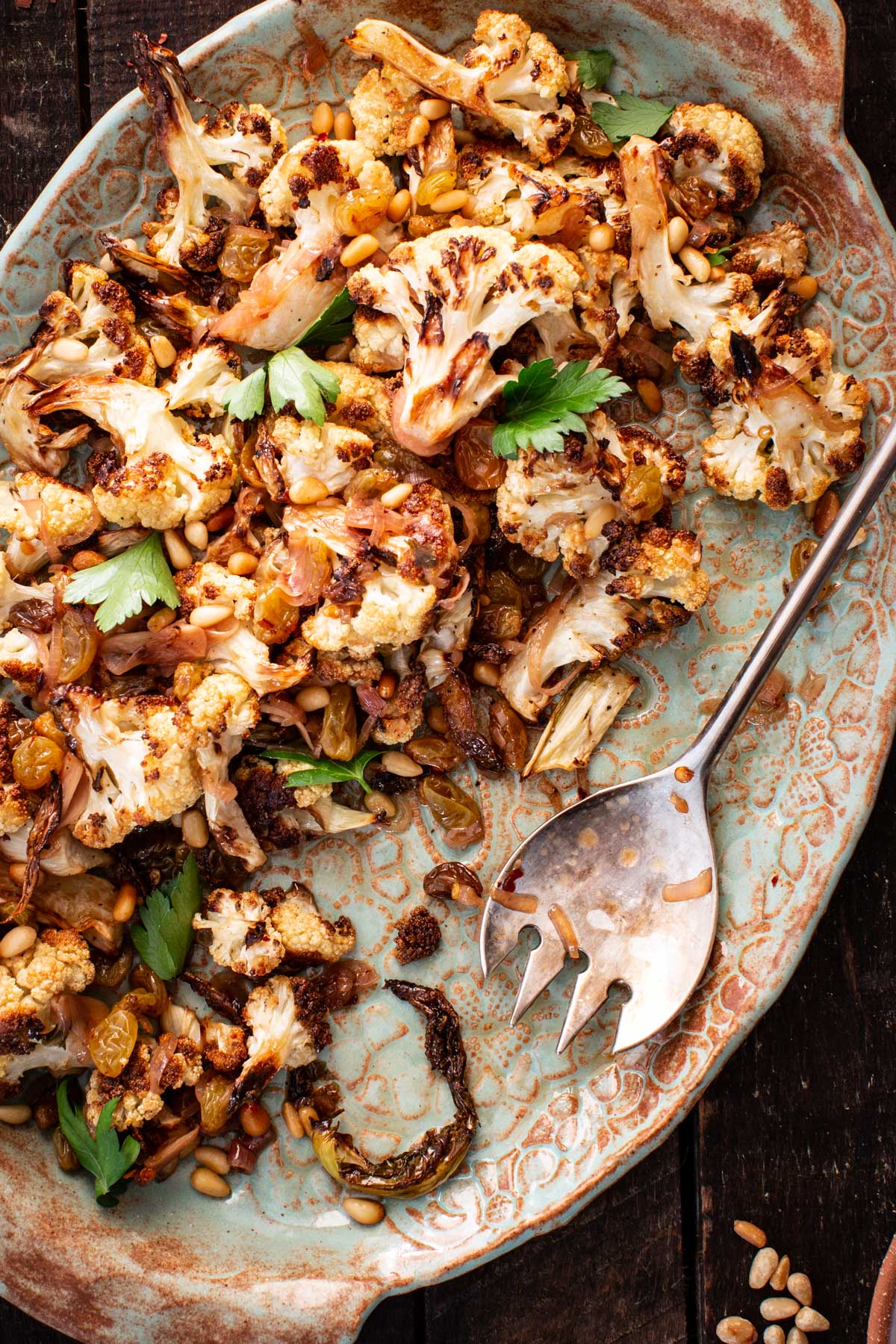 Roasted cauliflower agrodolce tossed with golden raisins, toasted pine nuts, and parsley on a rustic ceramic platter with a serving spoon.