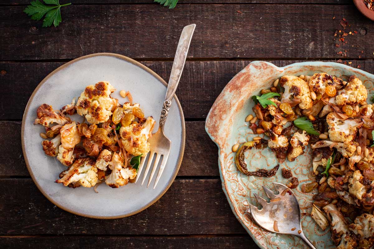 Roasted cauliflower agrodolce served on a small plate alongside the remaining platter, showing caramelized florets, raisins, pine nuts, and herbs on a wooden table.