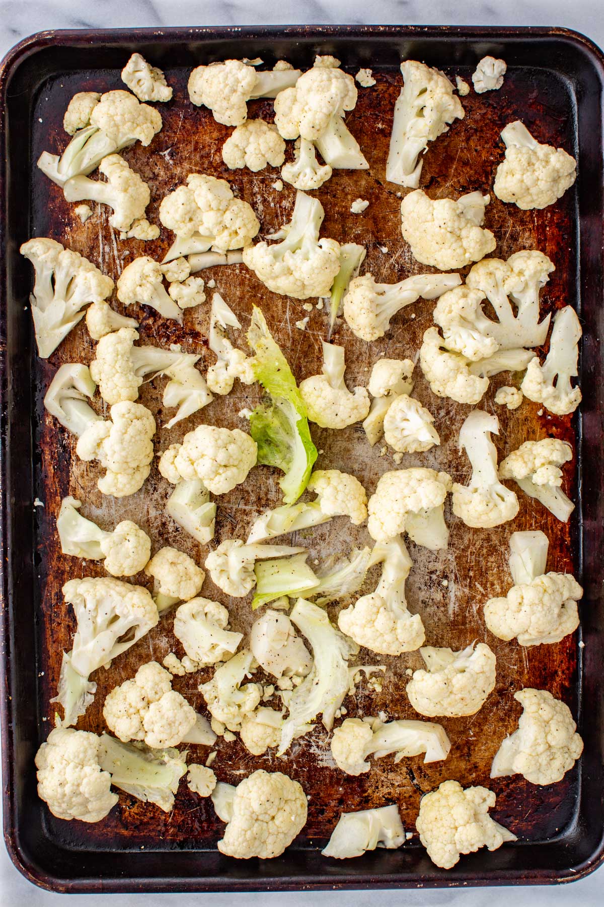 Cauliflower florets spread out on a well-used sheet pan, lightly seasoned and ready to be roasted.