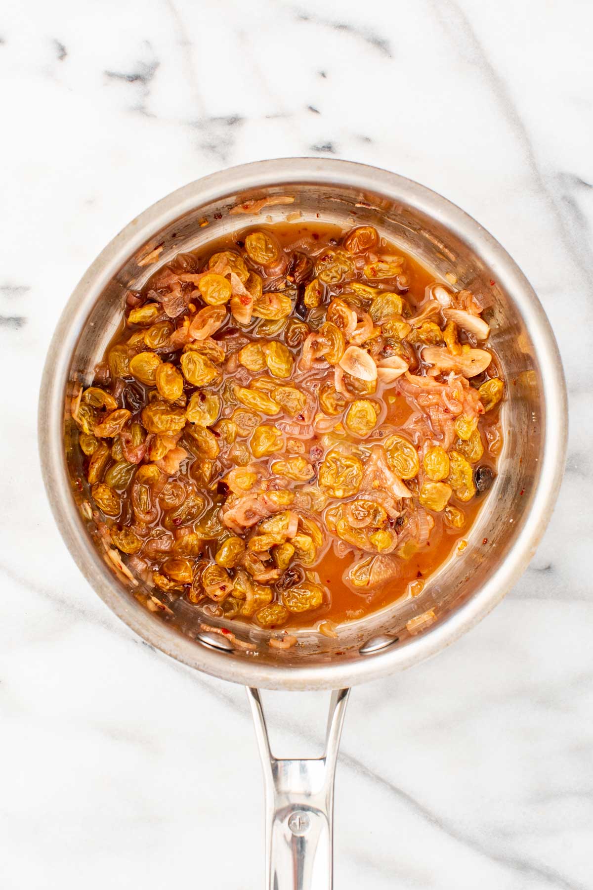 Golden raisins simmering with shallots, vinegar, and honey in a saucepan to create a sweet and sour agrodolce sauce.
