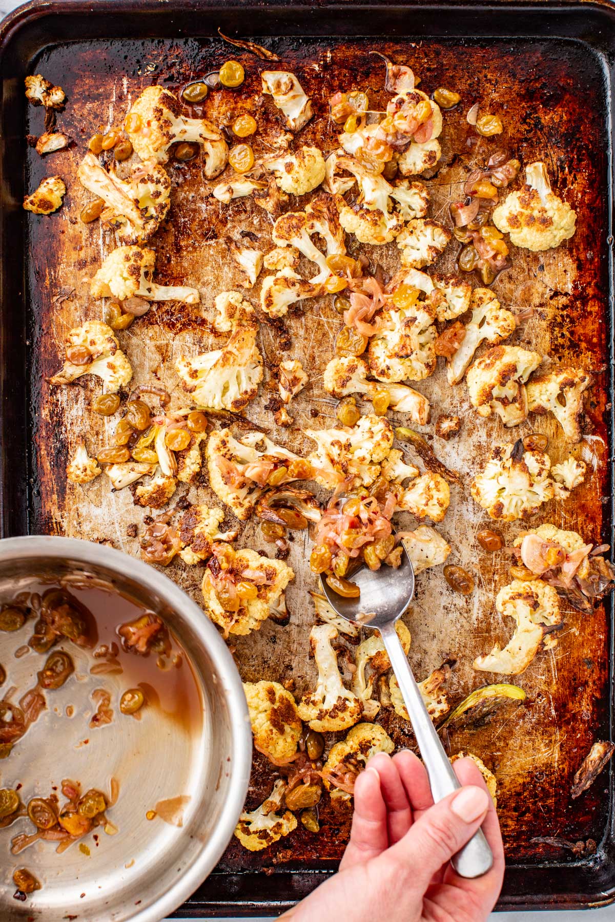 Agrodolce sauce being spooned over roasted cauliflower florets on a sheet pan.