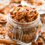 A glass jar filled with chunky homemade chai granola, surrounded by scattered granola clusters and cinnamon sticks on a light surface.