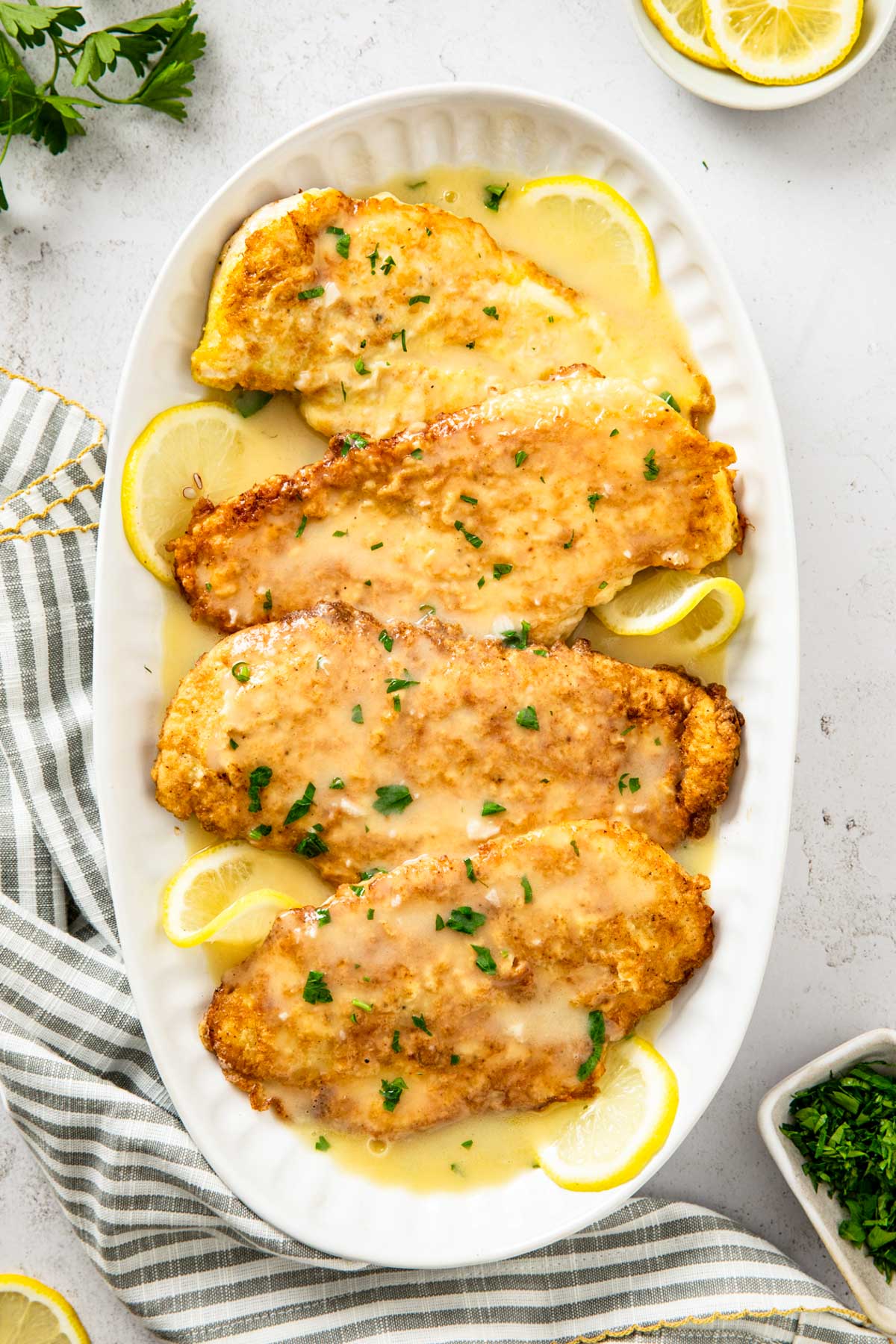 Chicken francaise arranged on an oval white platter with lemon slices, spooned with lemon butter sauce and finished with chopped parsley.