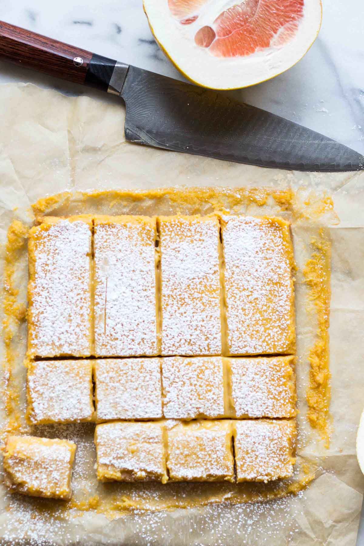 Overhead view of cut grapefruit bars dusted with powdered sugar, with a chef's knife and halved grapefruit on a marble surface.