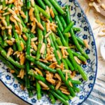 Buttery green beans tossed with toasted almonds and garlic, served on a blue and white oval platter with lemon on the side.