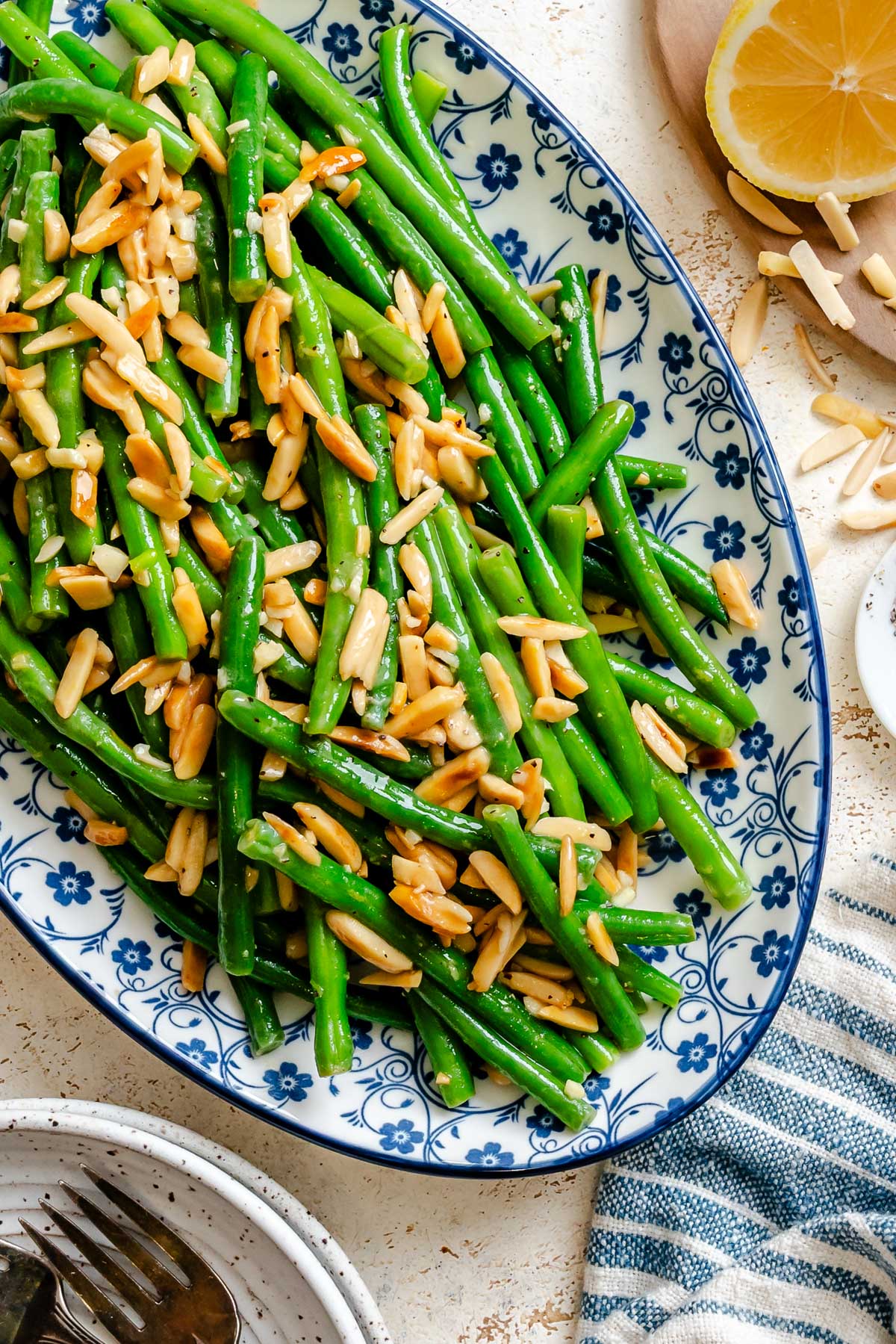 Buttery green beans tossed with toasted almonds and garlic, served on a blue and white oval platter with lemon on the side.