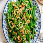 Overhead view of green beans amandine arranged on a patterned serving platter, with golden toasted almonds scattered throughout.