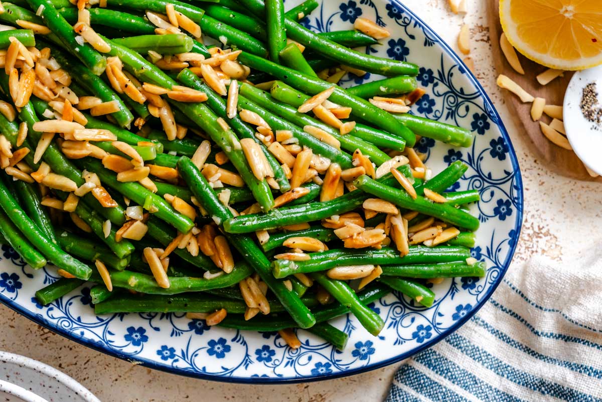 Close up of glossy green beans coated in butter with toasted almond slivers and bits of garlic on a blue floral plate.