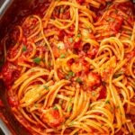 Overhead view of spaghetti tossed with lobster fra diavolo sauce in a wide pan, with chunks of lobster coated in a rich red tomato sauce and flecks of parsley.