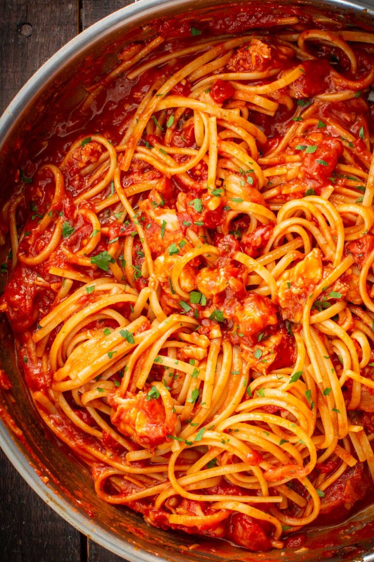 Overhead view of spaghetti tossed with lobster fra diavolo sauce in a wide pan, with chunks of lobster coated in a rich red tomato sauce and flecks of parsley.