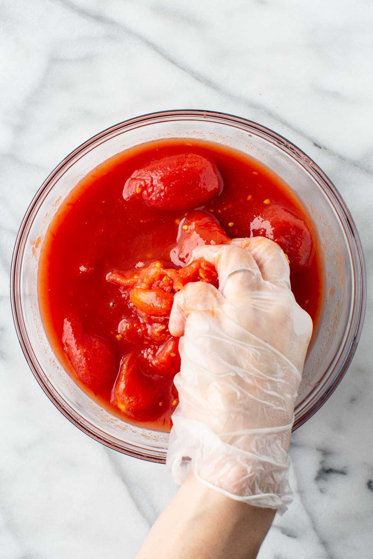 Gloved hand crushing whole peeled tomatoes in a glass bowl to make a rustic pizza sauce.