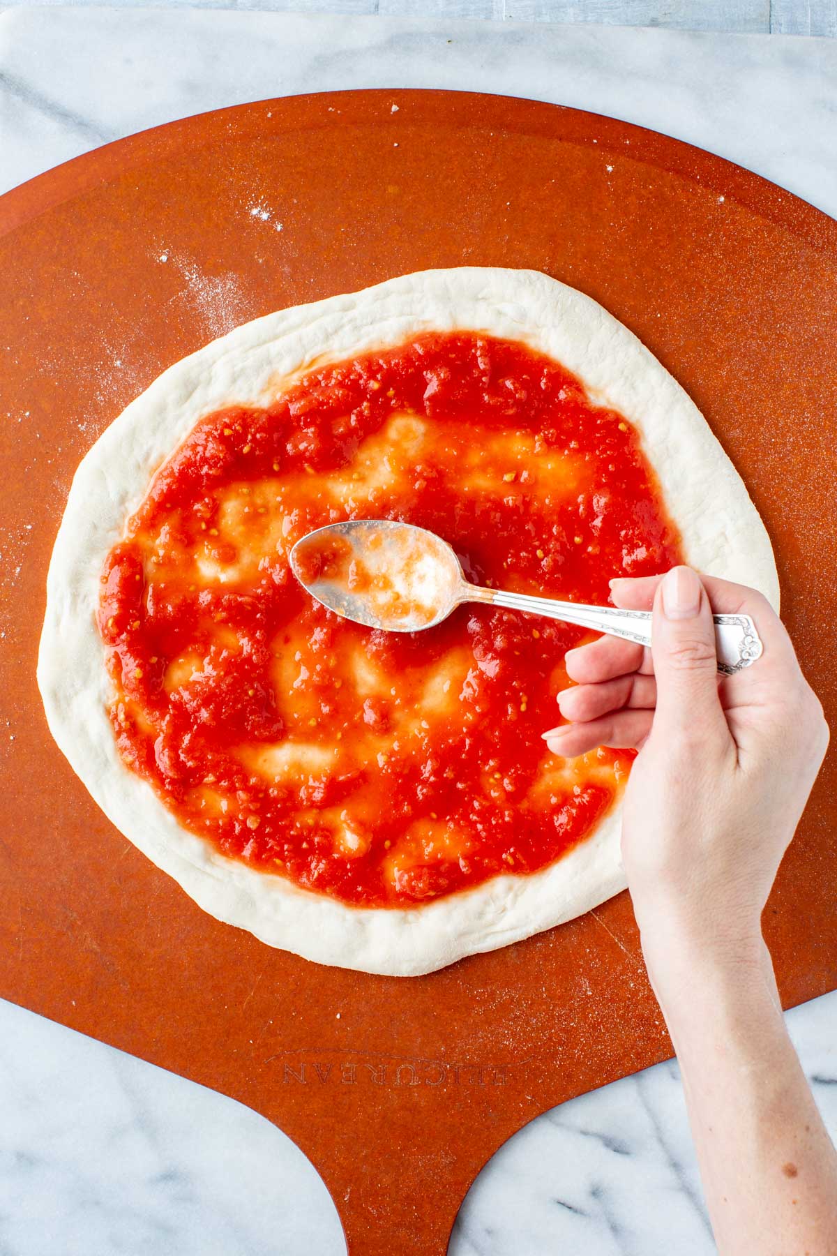 Spoon spreading crushed tomato sauce over stretched pizza dough on a wooden pizza peel.