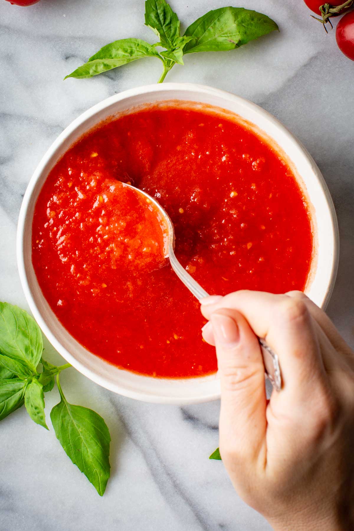 Spoon lifting bright red pizza sauce from a white bowl, garnished with fresh basil leaves.