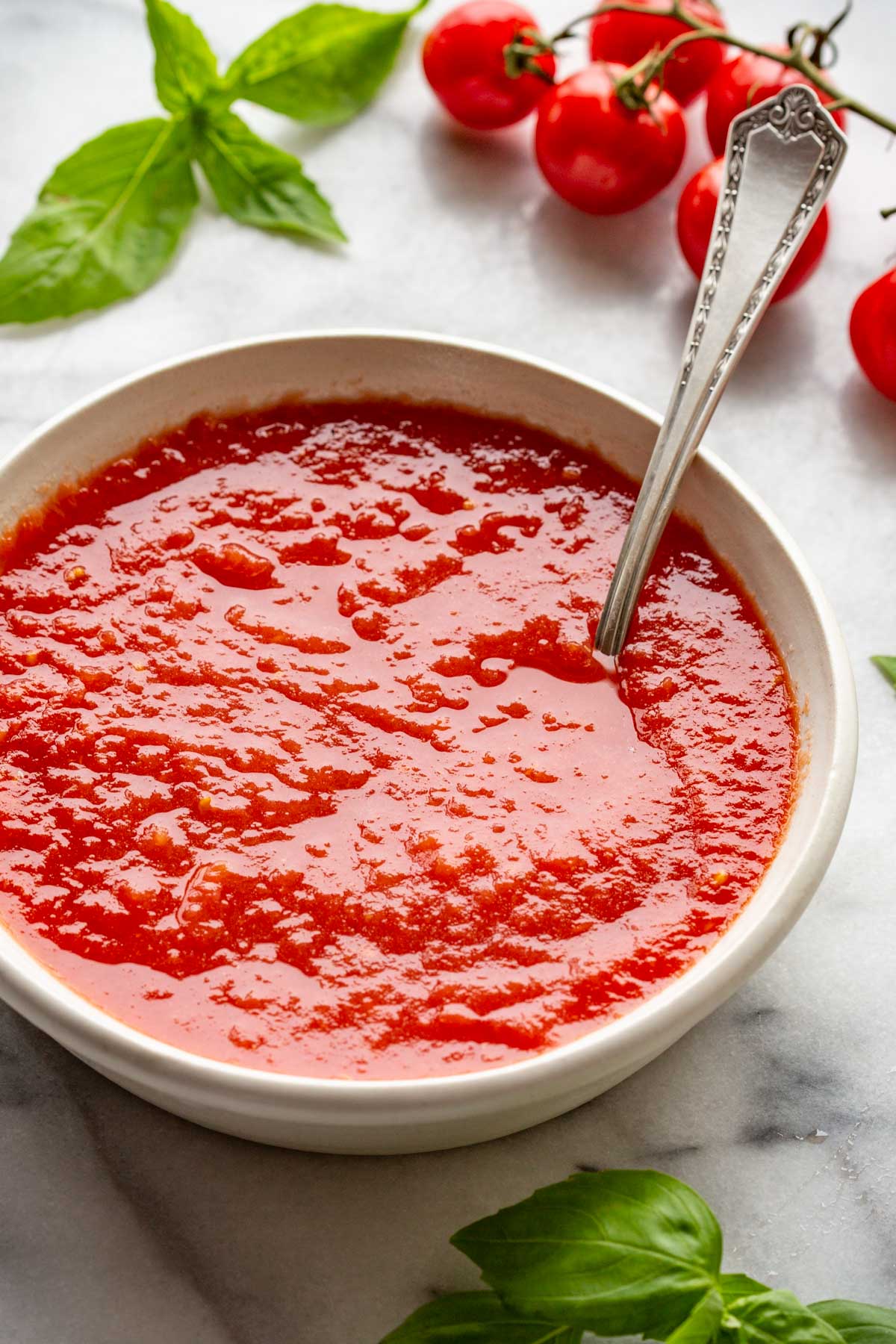 Smooth, vibrant red pizza sauce in a shallow white bowl with basil leaves and cherry tomatoes nearby.