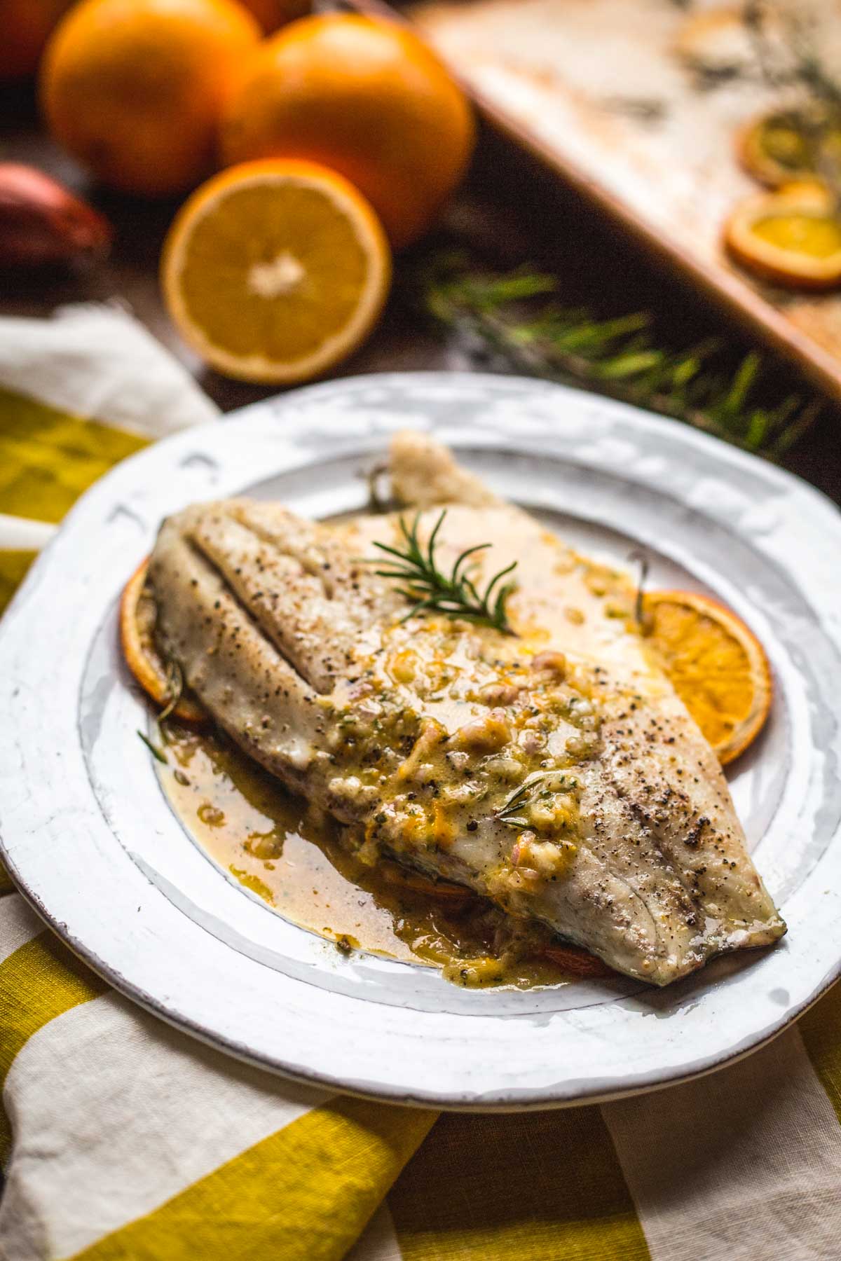 Roasted fish fillet served on a white plate with orange slices and rosemary, spooned with pan juices and citrusy sauce.