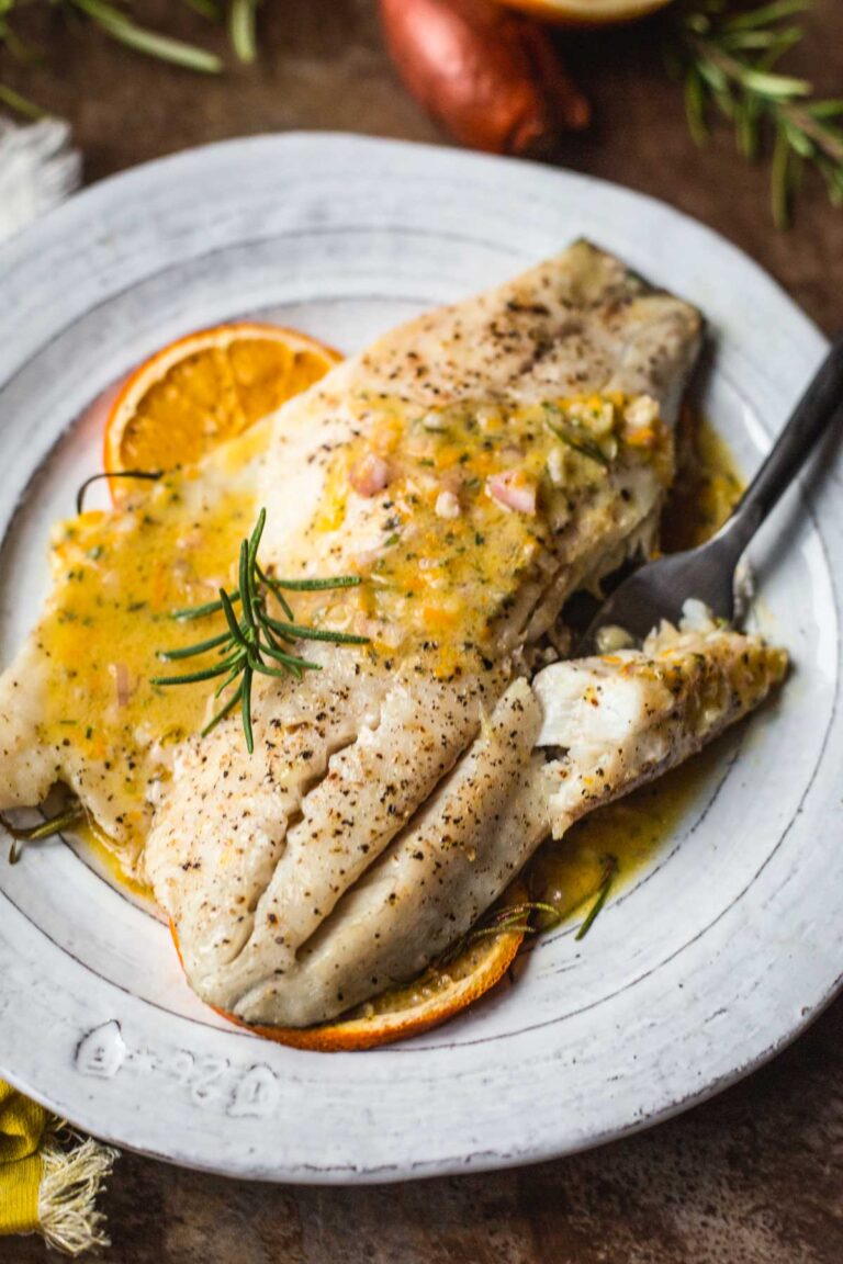 Close up of flaky roasted fish topped with orange rosemary pan sauce, cracked black pepper and fresh rosemary.