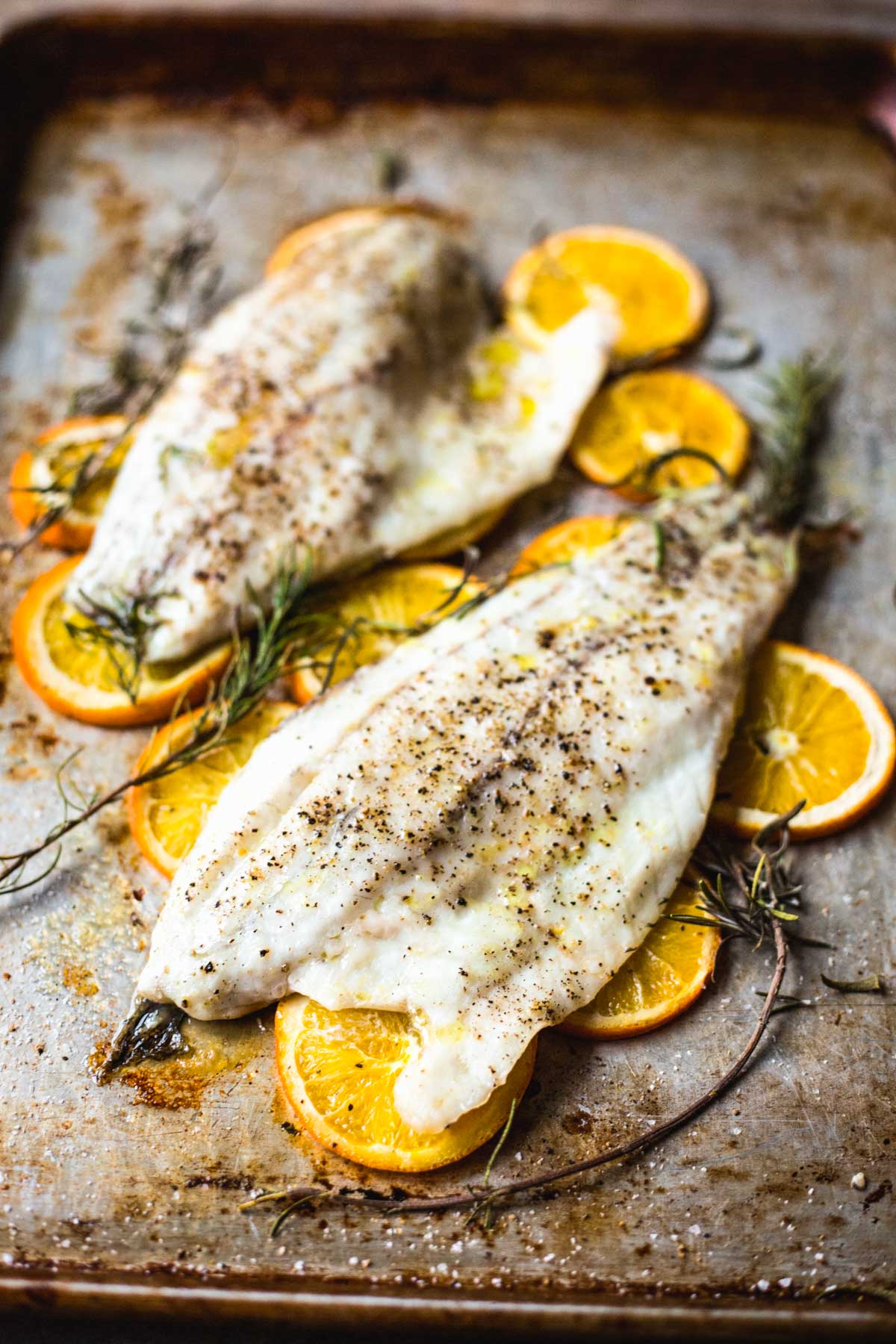 Two roasted fish fillets baking on a sheet pan with caramelized orange slices, rosemary sprigs and olive oil.